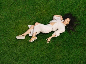 Rina Sawayama lying on lush green grass wearing light-coloured apparel and sandals for the Ugg Spring 2026 Golden collection campaign.