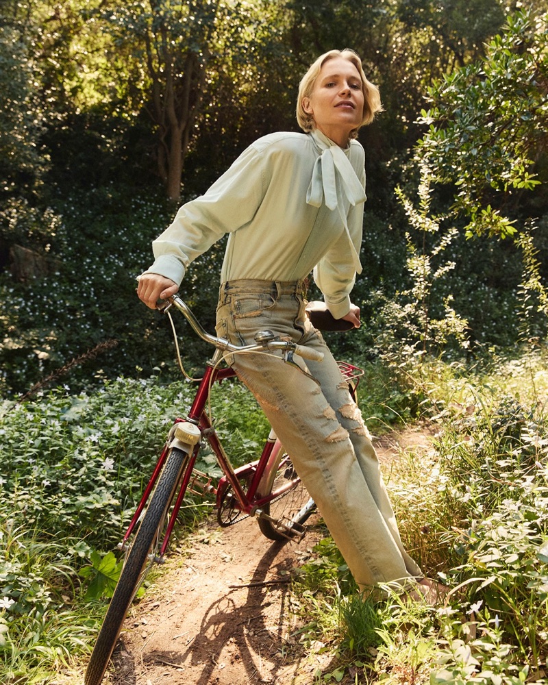 Model Steffi Cook wearing a mint green bow blouse and distressed jeans while posing on a vintage red bicycle in a sunlit woodland.