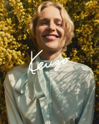 Model with blonde hair smiling in a light blue blouse against a dense background of yellow flowers