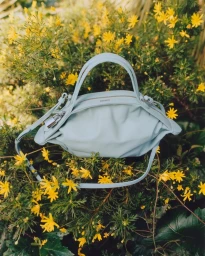 Light blue leather handbag nestled amongst green foliage and small yellow flowers outdoors.