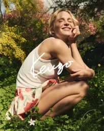 Blonde woman wearing white tank top and floral skirt crouching outdoors among blooming spring flowers for Kenzo Spring Flowers project