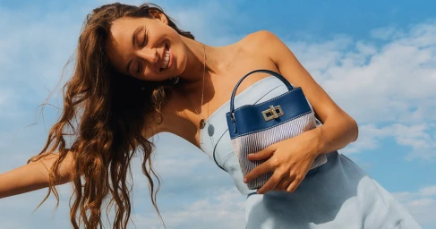 Smiling woman in light blue dress holding a blue and white woven handbag against a clear blue sky