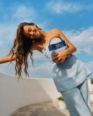 Model wearing a light blue strapless top and trousers holding a blue and white handbag against a clear sky background