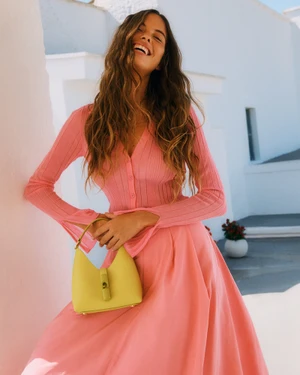 Smiling model in a pink ribbed dress holding a yellow handbag against white buildings