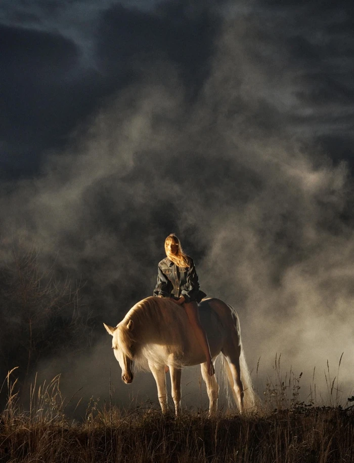 Model riding a white horse under a dramatic dark sky with atmospheric smoke or dust swirls