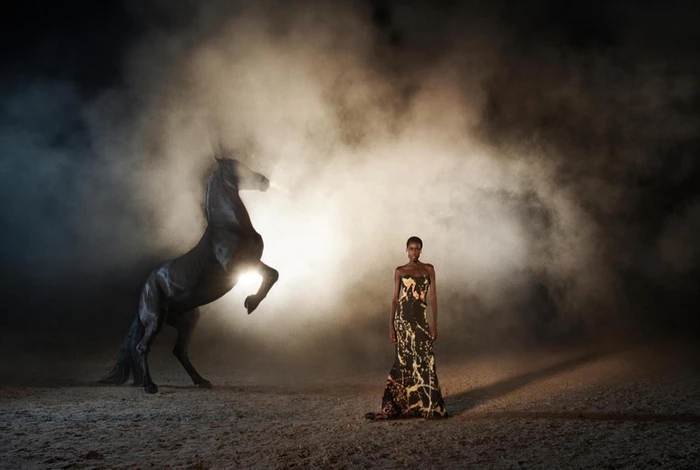 Model standing in a long printed dress next to a rearing black horse in a dusty arena filled with smoke and backlight.