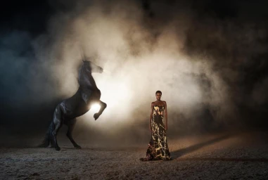 Model standing in a long printed dress next to a rearing black horse in a dusty arena filled with smoke and backlight.