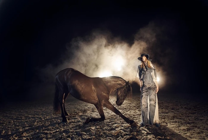 Model wearing denim outfit and cowboy hat standing beside a dark horse kneeling in a dusty arena at night with dramatic backlighting.