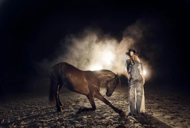 Model wearing denim outfit and cowboy hat standing beside a dark horse kneeling in a dusty arena at night with dramatic backlighting.