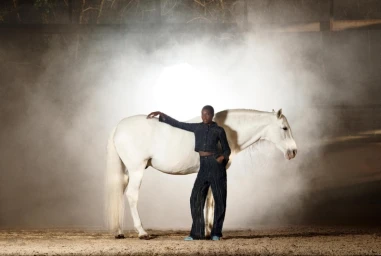 Model posing with a white horse in a dusty arena filled with atmospheric stage smoke under bright backlighting for Zara Western Arena.