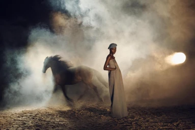 Model standing in a long dress and cap on dusty ground next to a running horse amid heavy smoke and a bright light source