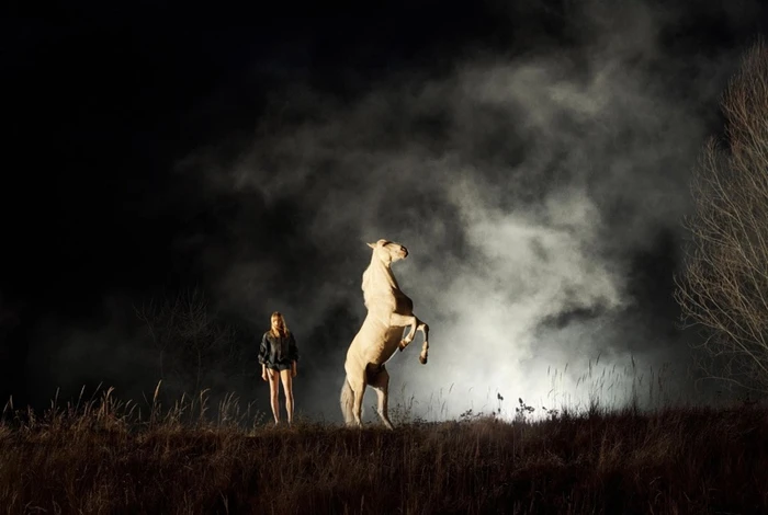 A woman stands beside a rearing white horse at night under smoky skies and dramatic lighting for a Zara campaign.