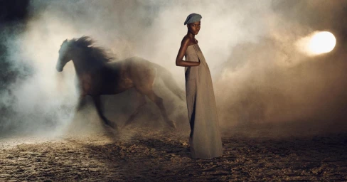 Model standing on a sandy ground wearing a long beige dress and beret with a running horse and smoke in the background.