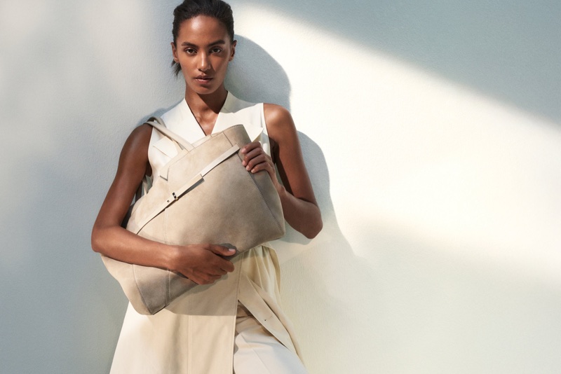 Model Sacha Quenby holding a beige suede tote bag while wearing a cream sleeveless top against a sunlit wall.