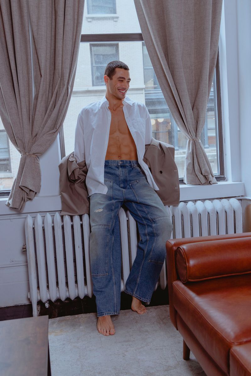 Man in unbuttoned shirt and jeans poses barefoot by window with curtains and radiator in indoor setting with leather couch.