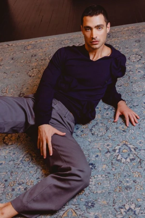 Adult male lying on an ornate carpet wearing grey trousers and a dark blue, long-sleeved shirt with a distinct collar.