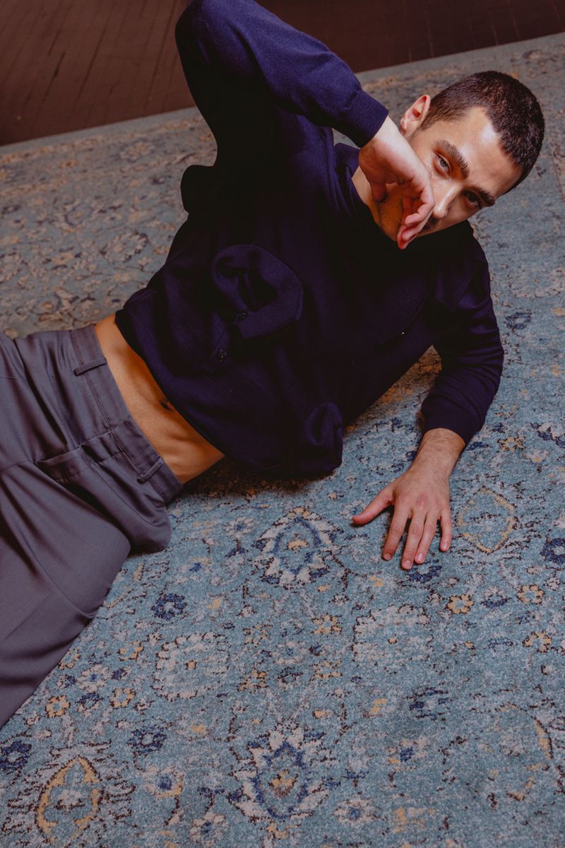 A young adult man lies on an ornate patterned area rug, wearing a dark blue sweater and grey pants, in an indoor setting.