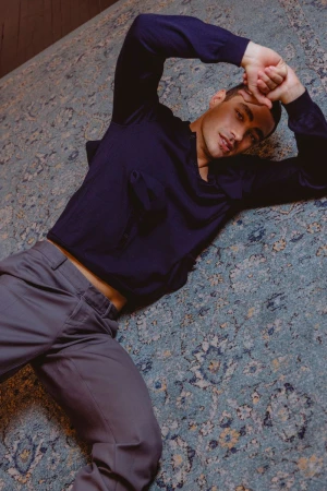 Attractive adult male lies on a patterned rug indoors, wearing a long-sleeve shirt and dress pants with natural lighting and soft focus.