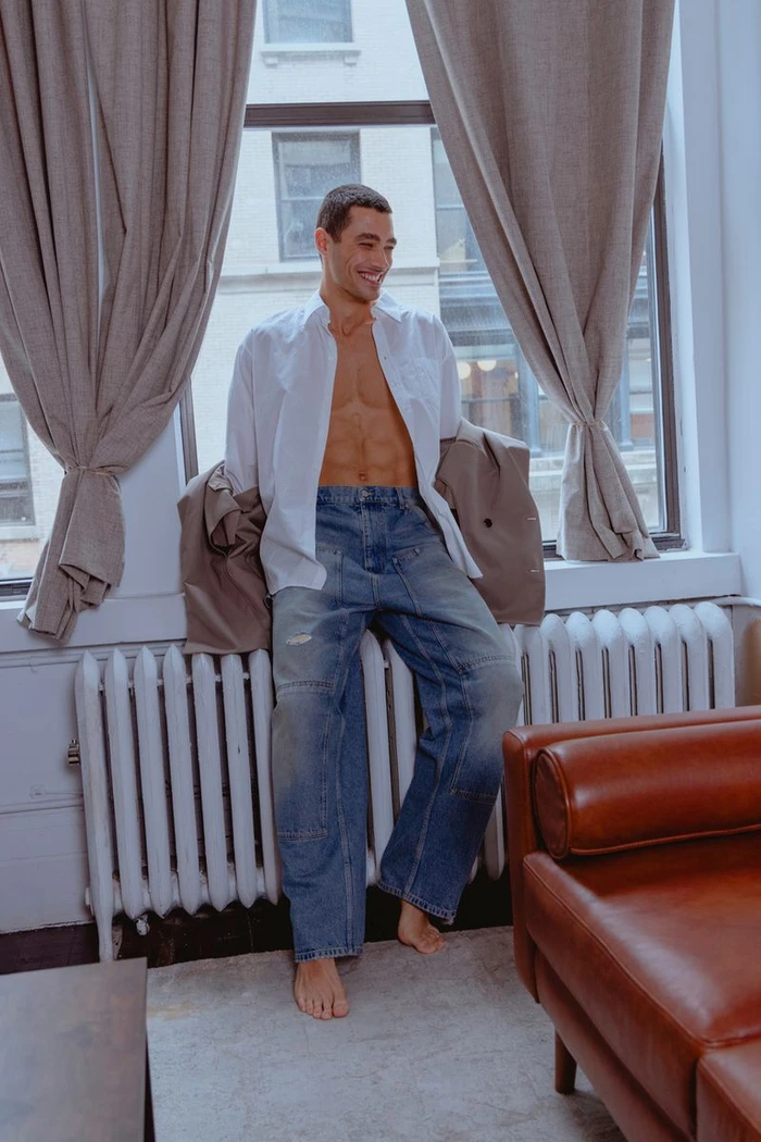 A smiling man wearing an unbuttoned white shirt and wide-leg blue jeans leans against a radiator beside draped window curtains.