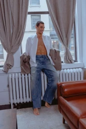 A smiling man wearing an unbuttoned white shirt and wide-leg blue jeans leans against a radiator beside draped window curtains.