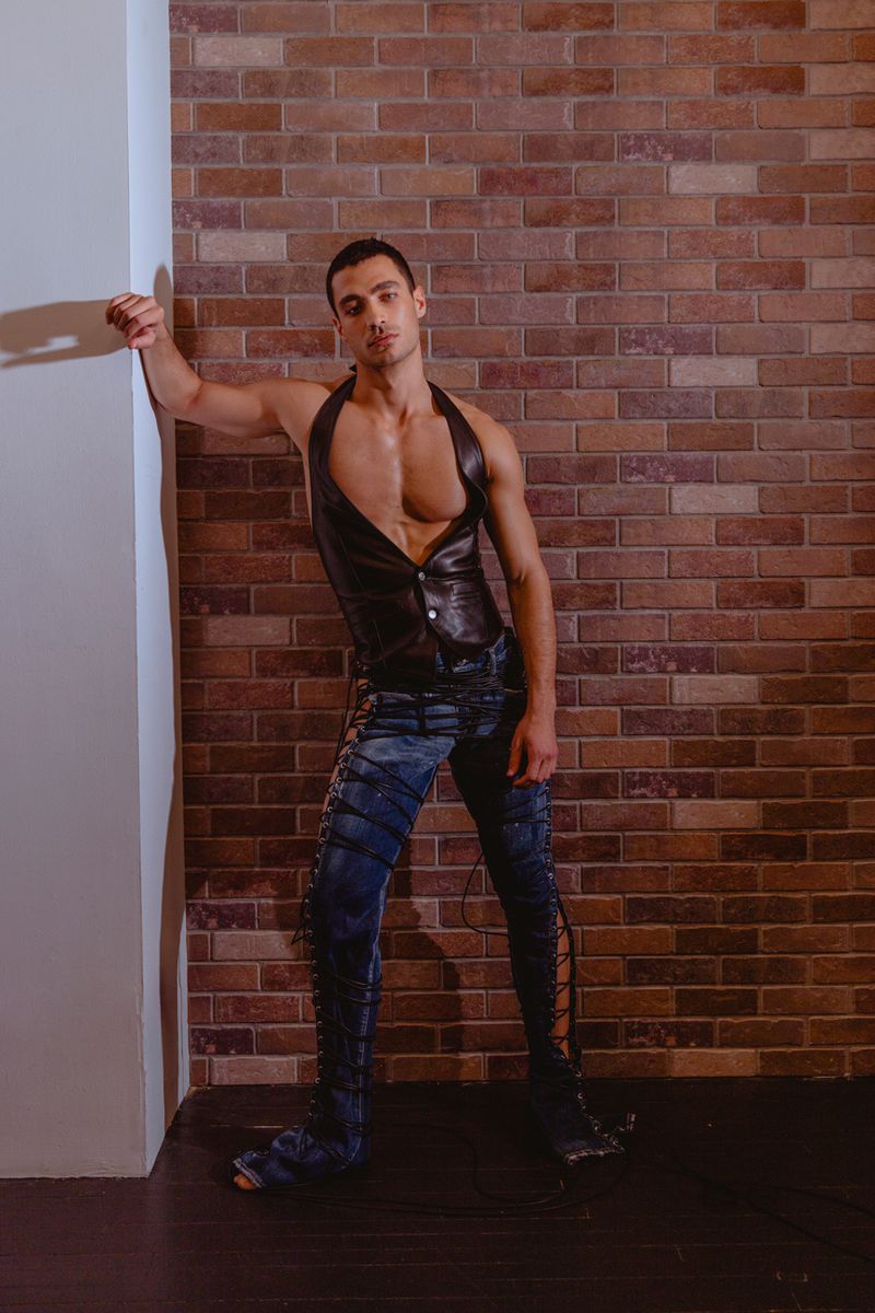 Male model in a black leather halter vest and lace-up denim pants posing against an exposed brick wall.