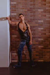 Male model in a black leather halter vest and lace-up denim pants posing against an exposed brick wall.