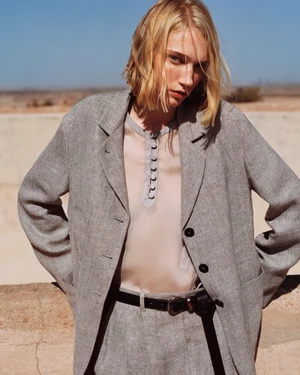Model with blonde bob wearing a grey blazer over a light blouse with dark buttons against an outdoor arid backdrop.