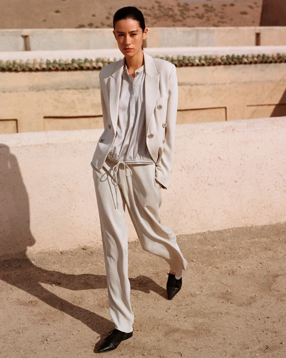 Model wearing a light-coloured blazer and trousers standing outdoors against a warm earth-toned architectural wall.