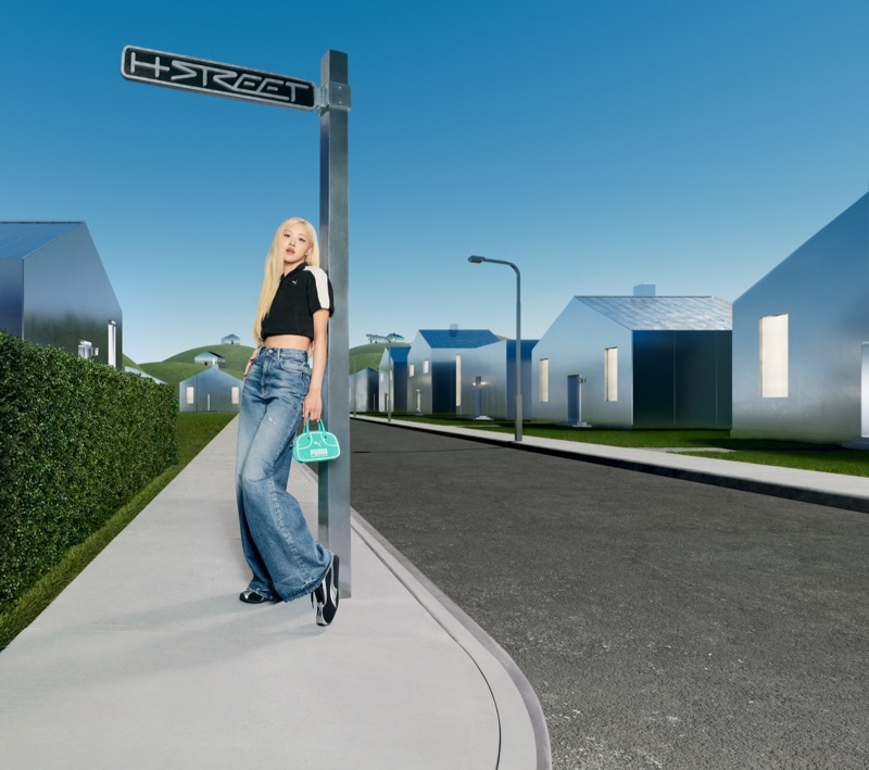ROSÉ poses in a black crop top and wide-leg denim jeans leaning on an H-Street signpost on a surreal street.