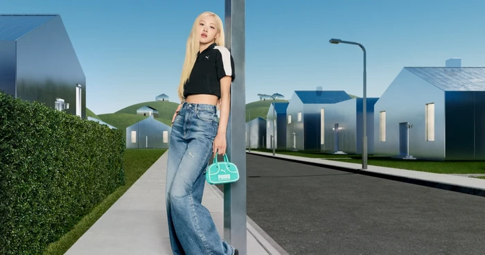 ROSÉ leaning against a pole wearing a black top and blue jeans, holding a teal Puma handbag in a surreal suburban neighborhood setting.