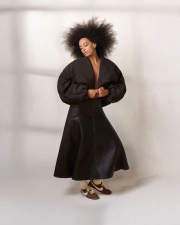 Solange Knowles in a black cropped puff jacket, long flared black skirt, and brown Nike sneakers against a studio backdrop.