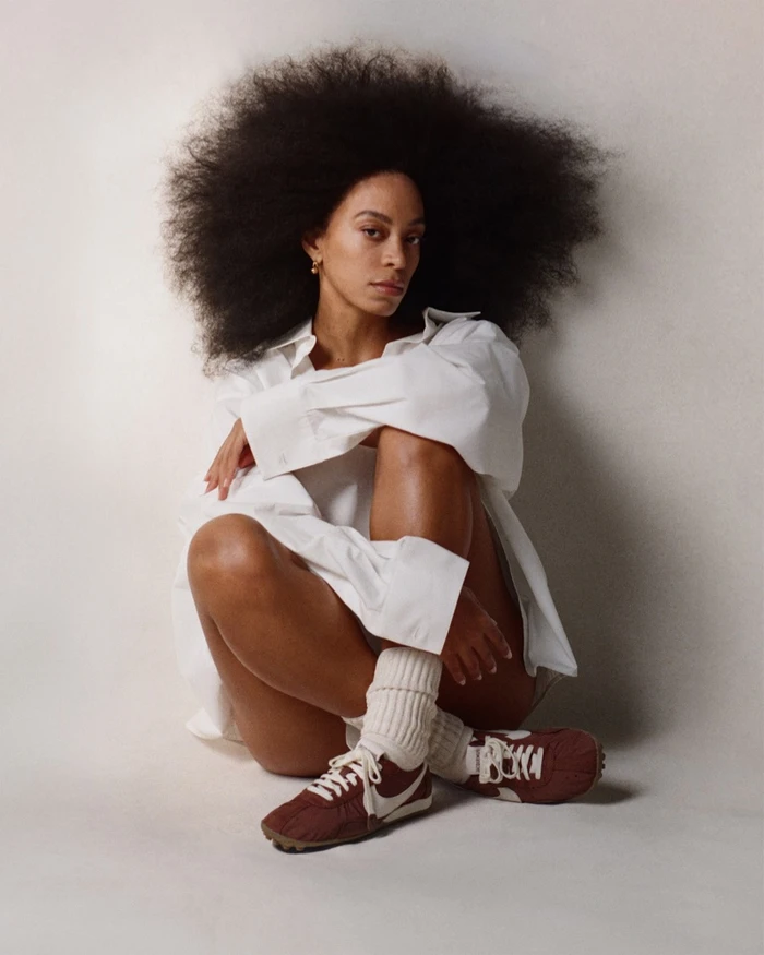 Woman with voluminous afro hairstyle wearing a white shirt and brown sneakers sitting against a white background