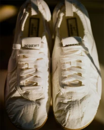 Close-up front view of white Nike Moon Shoe sneakers featuring Jacquemus branding on the tongue under warm lighting.