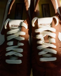 Close-up front view of brown Nike Moon Shoe sneakers with white laces and Jacquemus branding on the tongue under warm lighting.