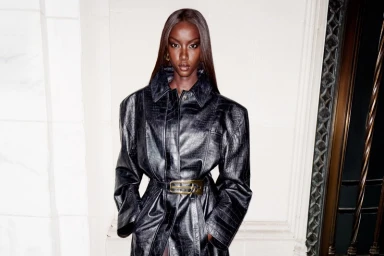 Anok Yai wearing a black textured leather belted coat standing before architectural stonework for Revolve Los Angeles.
