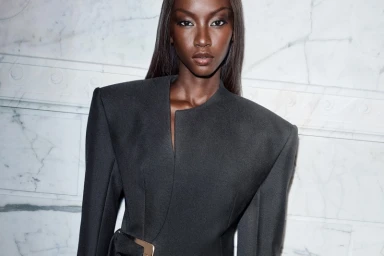 Anok Yai wearing a structured dark coat with strong shoulders against a white marble wall for Revolve Los Angeles.