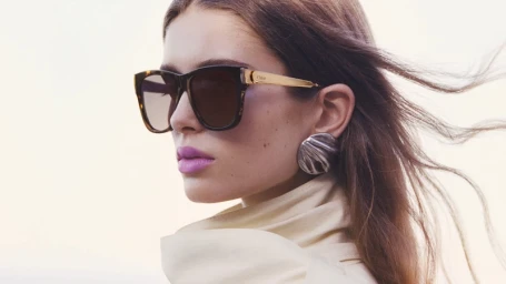Close-up portrait of a woman wearing tortoiseshell sunglasses and large silver earrings against an outdoor sky background