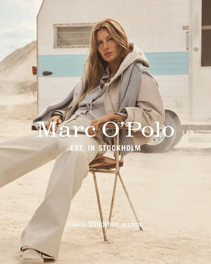 Gisele Bundchen seated on a chair outdoors wearing a beige jacket and light trousers with a caravan in the background