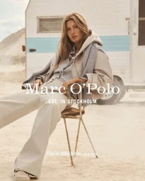 Gisele Bundchen seated on a chair outdoors wearing a beige jacket and light trousers with a caravan in the background