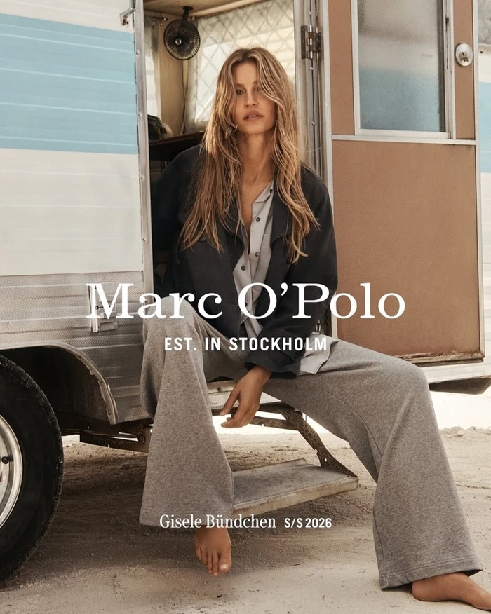 Gisele Bündchen seated in the doorway of a vintage camper van wearing a dark jacket and grey trousers for Marc O'Polo Spring 2026 campaign