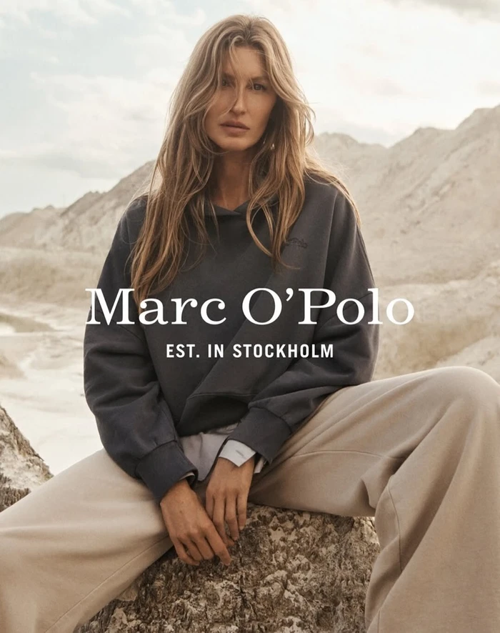 Gisele Bundchen wearing a dark grey hoodie and light trousers outdoors against a sandy terrain background.