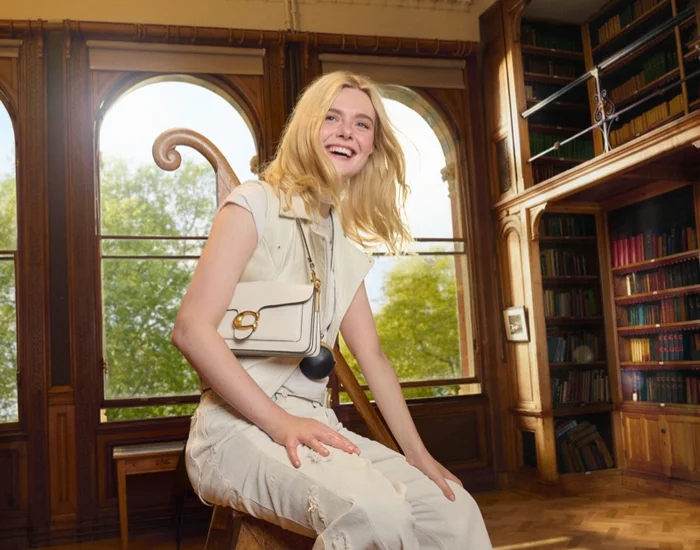 Elle Fanning sitting in a wood-paneled library wearing cream clothing and holding a white Coach shoulder bag