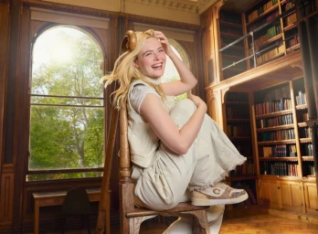 Elle Fanning sitting on a large wooden chair in a library wearing off-white casual apparel and sneakers for Coach