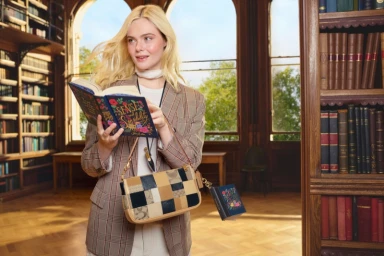 Elle Fanning holding a book in a library wearing a plaid blazer and carrying a patchwork leather shoulder bag.