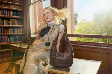 Elle Fanning sitting in a wood-paneled library holding a brown quilted leather shoulder bag for Coach Explore Your Story