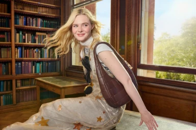 Elle Fanning holding a dark brown leather shoulder bag in a library with tall wooden bookshelves and a large window.