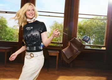 Elle Fanning wearing a Coach graphic t-shirt and white trousers holding a brown leather shoulder bag near large windows