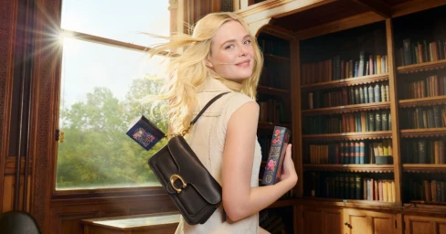 Elle Fanning holding a book in a library, wearing a beige sleeveless top and carrying a dark leather shoulder bag with a gold clasp.