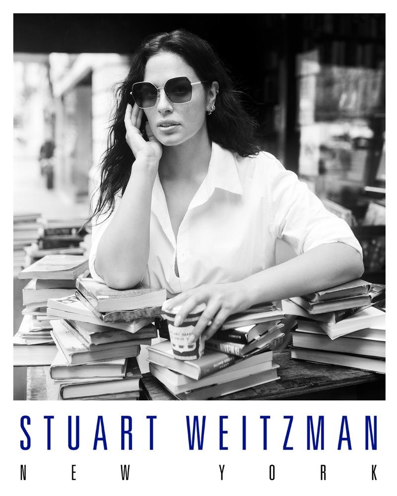 Ashley Graham wearing sunglasses and a white shirt leaning on stacks of books for Stuart Weitzman.