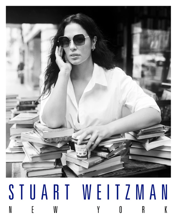 Ashley Graham wearing sunglasses and a white shirt leaning on stacks of books for Stuart Weitzman